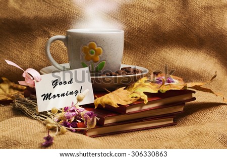 Tag with words good morning and the cup of coffee lying on the books with dry yellow maple leaves and everlasting flowers on sackcloth background