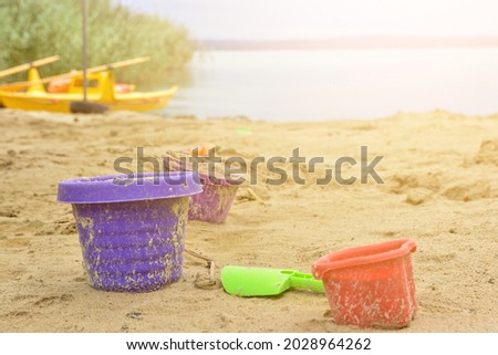 Colorful sandbox toys on a sandy lido beach with the blue water, reeds and a yellow boat in the background on a sunny summer day, children's leisure time activity, purple plastic bucket Foto stock © 