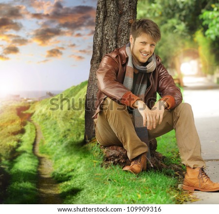 Young stylish happy man in autumn clothing leaning against tree next two path fading off in the distance