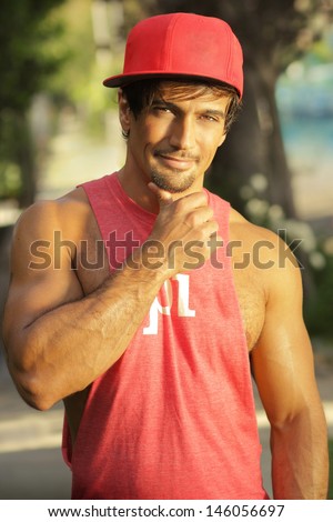 Cool young guy in cap and muscle shirt outdoors with playful flirty smirk