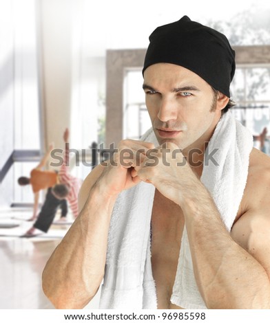 Portrait of a serious muscular male fitness model inside gym with fists raised to box wearing a hat and towel with blurred background of active sports gym