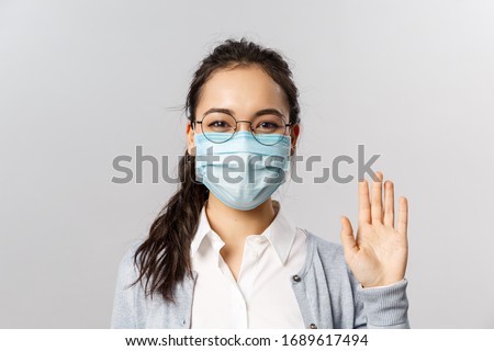 Covid19, virus, health and medicine concept. Portrait of friendly young girl in medical face mask saying hi, hello waving hand and grinning with eyes, keep safe during pandemia, quarantine
