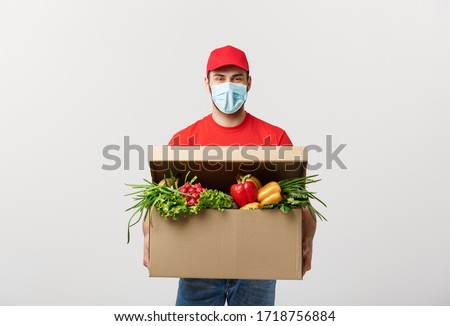 Delivery Concept: Handsome Caucasian grocery delivery courier man in red uniform and face mask with grocery box with fresh fruit and vegetable