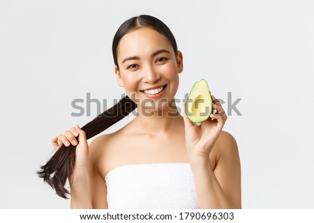 Beauty, personal care, spa and skincare concept. Close-up of happy good-looking asian woman in bath towel, showing hair and avocado, promo of shampoo, haircare masks or conditioner
