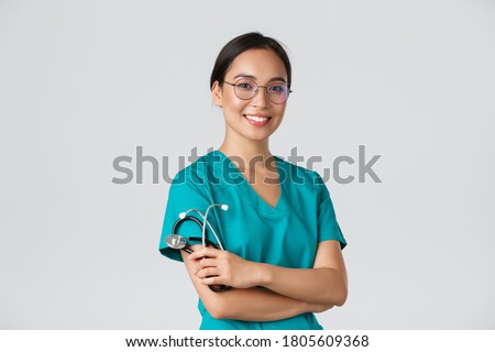 Covid-19, coronavirus disease, healthcare workers concept. Professional good-looking asian doctor, medical worker in glasses and scrubs, cross arms and smiling, white background