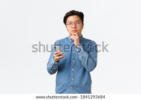 Thoughtful creative asian man in glasses thinking while making post on social media, looking away, pondering or making decision, holding smartphone, choosing something in internet