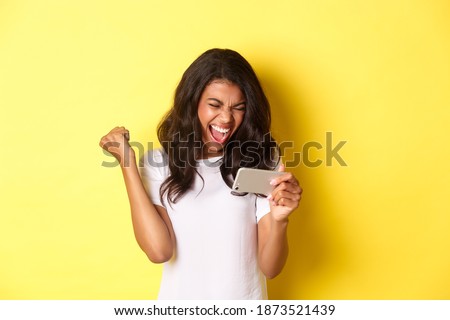 Portrait of cute african-american girl winning in smartphone game, shouting for joy, making fist pump gesture and looking at phone screen, standing over yellow background ストックフォト © 