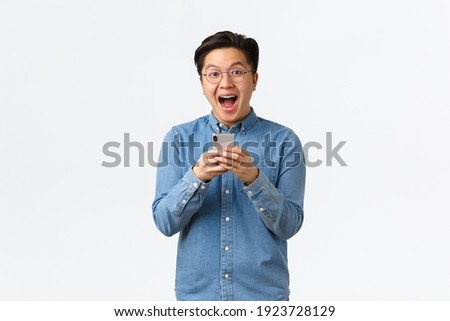 Excited happy asian man reacting cheerful at awesome news read online, holding mobile phone and looking thrilled with event coming up. Guy download cool new app or game, white background
