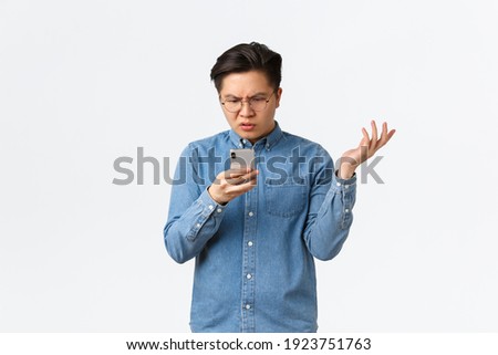 Frustrated and confused asian guy in glasses looking puzzled at mobile phone screen, raising hand up questioned, cant understand what happening online, standing white background perplexed Foto stock © 