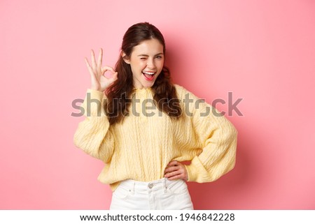 Cheeky young woman winking, showing okay sign, give her approval, like and approve good thing. Girl make OK gesture to give permission, say yes, standing over pink background