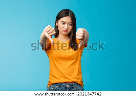 Upset gloomy asian girl with dark haircut frowning sulking sad, disappointed, show thumbs-down grimacing, give negative opinion, judging bad uninteresting movie, stand blue background