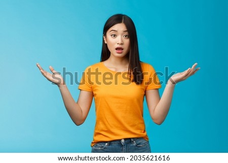 Questioned unaware young asian girl with dark short haircut shrugging hands spread sideways, look confused open mouth uncertain, being clueless, stand blue background wear yellow t-shirt