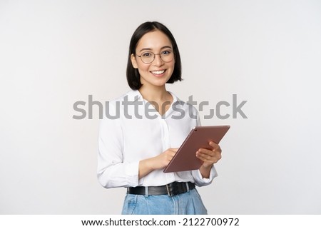 Image of young asian woman, company worker in glasses, smiling and holding digital tablet, standing over white background Foto d'archivio © 
