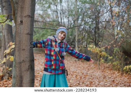 happy young girl with stretched arms staying behind the tree in the park in autumn