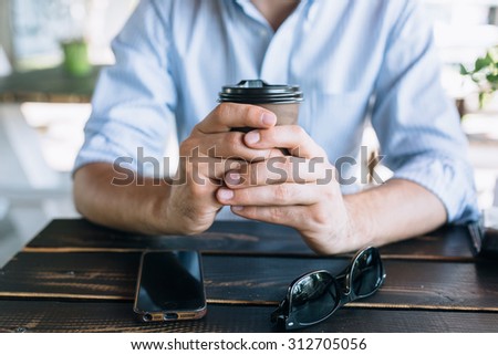 Young stylish man sitting with a cup of coffee in the cafe outdoors. Mobile phone and sunglasses in front of the cup.