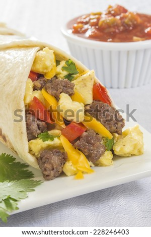 close up of a hot fresh breakfast burrito with eggs,sausage,cheese,tomatoes,cilantro, and salsa on the side \