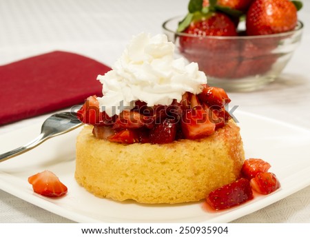 Bright red strawberries topped with syrup and whipped cream on a yellow shortcake. Served on a square plate set on a white table cloth. Bowl of strawberries in the background.\