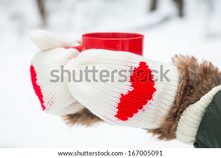 Hands in warm mittens with hearts holding red cup close-up