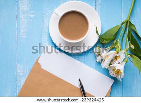 Cup of coffee with milk, blank paper in the envelope, pen and alstroemeria flowers on the blue colored wooden table, top view