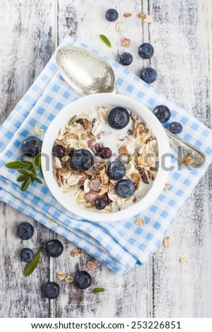Light breakfast setup with bowl of muesli, metal spoon and fresh blueberries on blue checkered cloth over white painted grunge style wooden table.