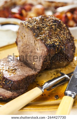 Carved roast beef joint with carving knife and fork on oval plate over rustic wooden table with pan of roasted vegetables and jug of sauce on the background