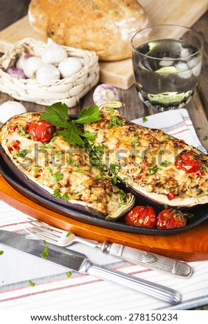 Aubergine halves stuffed with vegetables and cheese on oval cast iron pan with garlic, tomatoes, bread and glass of water over the rustic wooden table