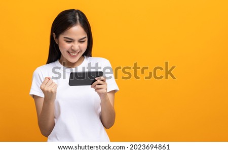 Excited Beautiful young asian women play mobile game and standing on isolated yellow background. Playing game on smartphone winning victory moment. Very enjoy and fun relax time