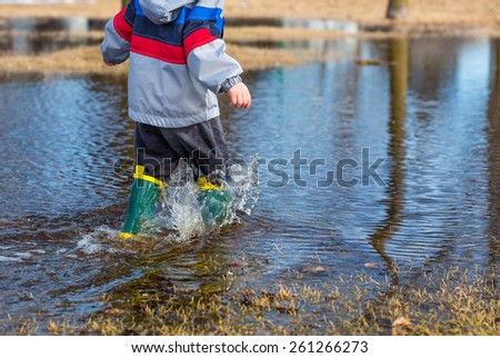 Very wet little boy running through puddles in the grass wearing rain boots.