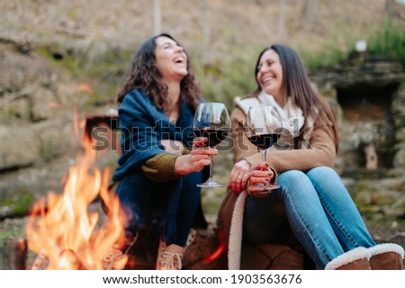 young happy women laughing, holding glass of red wine. Females warming next to the fire. Campfire, outdoors activities concept.