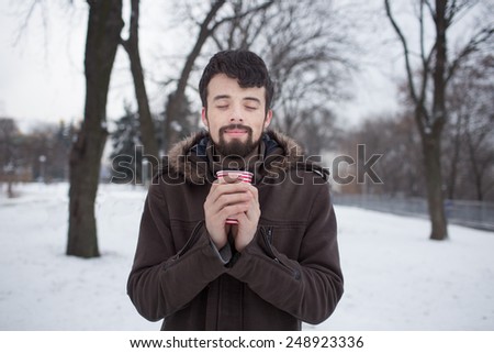 one black-haired, bearded man in a brown Platja is a snowy winter park and holding a cup cocoa or tea. Hispanic cute kid hands warm cup of coffee.