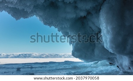 In the cave, rows of fancy blue icicles, similar to lace, hang from the vault of the grotto. The walls are icy. Ahead - a clear azure sky, a snow-covered mountain range, a frozen lake. Baikal.  商業照片 © 