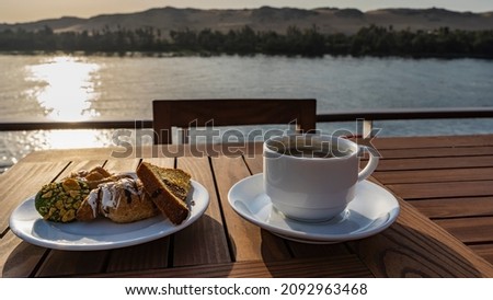 There is a wooden table on the upper deck of the cruise ship. Cookies and cupcakes are laid out on a saucer. There's coffee in the cup. Soft background - the Nile River, sand dunes. Egypt. Foto stock © 