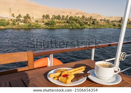 A saucer with cupcakes, pastries and a cup of coffee stands on a wooden table on the deck of a cruise liner. Behind the railing you can see the river, green vegetation and sand dunes against the sky. Foto stock © 