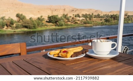 The deck of a cruise ship. On a wooden table there is a cup of coffee and a saucer with pastries, cupcakes. The background is a blue river, green vegetation on the shore, sand dunes. Egypt. Nile Foto stock © 