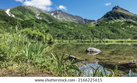 Quiet alpine lake. The rocks at the bottom are visible through the clear water. Lush green vegetation on the banks. A  mountain range against the blue sky. Kamchatka. Vachkazhets. Lake Tahkoloch 商業照片 © 