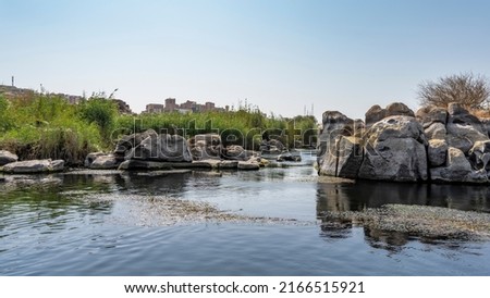 In a quiet river backwater there are piles of boulders, duckweed on the water. Green vegetation on the shore. Reflection. Clear blue sky. Egypt. Nile. Aswan 商業照片 © 