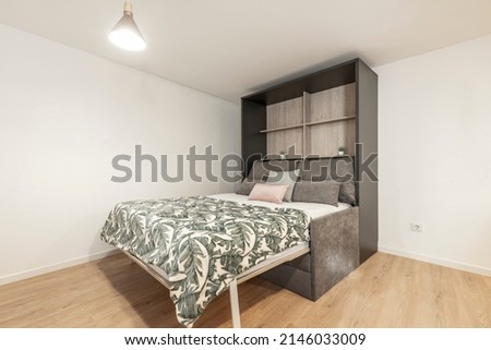 Folding bed inside dark gray wardrobe with cushions in various sizes and colors, white bedding, floral blanket, wooden shelves in vacation rental studio Foto stock © 
