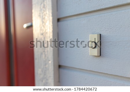 White plastic doorbell on white wooden facade with a red door in the background
