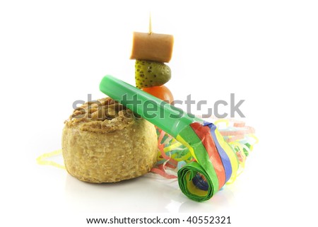 Small pork pie with party blower and cocktail stick containing hot dog sausage, gherkin and tomato with streamers on a reflective white background