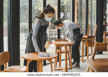 asian waitress staff wearing protection face mask in apron cleaning table with disinfectant spray for protect infection coronavirus (covid-19) in cafe coffee shop restaurant, hotel. new normal concept