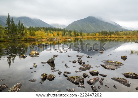 Kuelporr mountain with peak hidden in clouds reflected in shallow Polygonal northern taiga forest lake with lichen-covered rocks in foreground