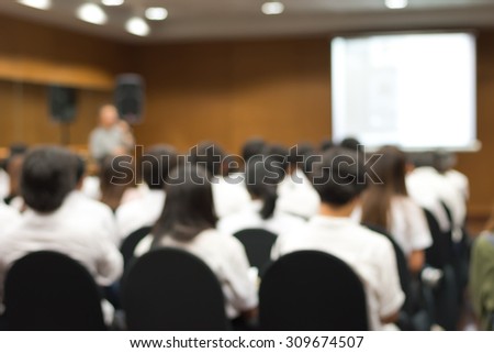 Blurred abstract background of university students sitting in a lecture room with teacher in front of the class with white projector slide screen: Blurry view from back of the classroom