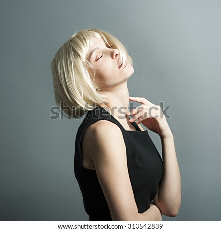 Beautiful skinny slender young blonde girl with a beautiful serene face and a long neck wearing a wig stands sideways in the studio in a strict black dress raising his hand up and his head back