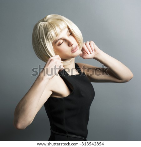 Beautiful skinny slender young blonde girl with a beautiful serene face and a long neck wearing a wig studio is in strict black dress hands up touched hair