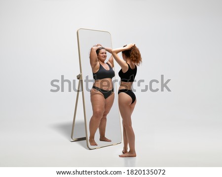 Young fit, slim woman looking at fat girl in mirror's reflection on white background. Thinking she's not enough sportive. Concept of healthy lifestyle, fitness, sport, nutrition and body positive. Imagine de stoc © 