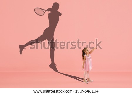 Childhood and dream about big and famous future. Conceptual image with girl and drawned shadow of female tennis player on coral pink background. Childhood, dreams, imagination, education concept.