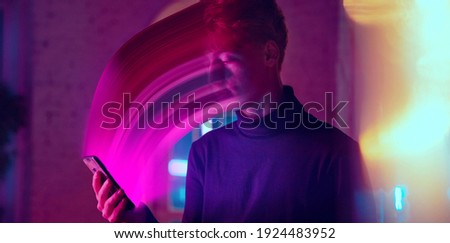 Cinematic portrait of stylish man in neon lighted interior using a smartphone. The face is smeared, sucked, absorbed into the phone. Concept of social network dependency, phone addiction. 商業照片 © 