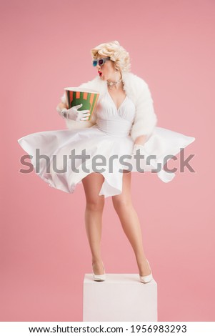 Cinema, movie time. Portrait of young blonde woman in white dress on coral pink background. Female model as an legendary actress. Pin up. Concept of comparison of eras, modern, fashion, beauty.