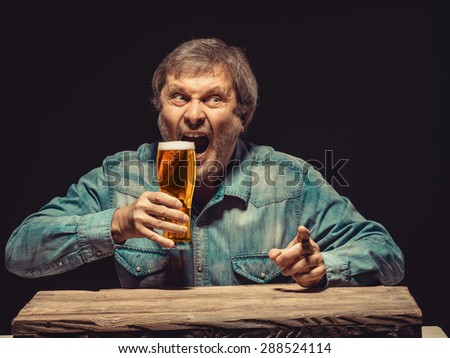 The front view of handsome screaming man as fan in denim shirt with glass of beer, sitting at the wooden table. Concept of emotional fan, interested showing something