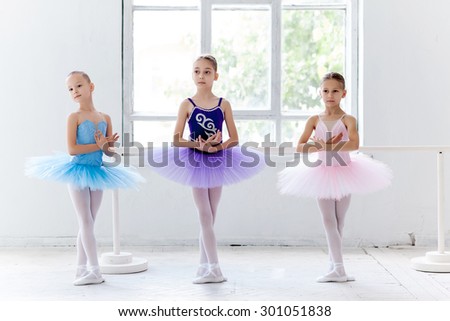 Three little ballet girls in multicolored tutu posing at ballet barre together in white studio